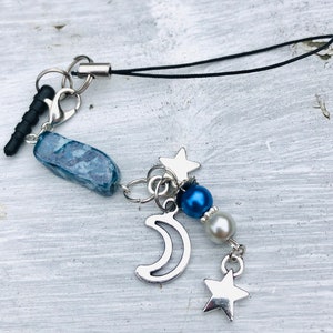 May include: A blue and white stone charm with a silver crescent moon, two silver stars, and blue and white beads hangs from a black phone strap.