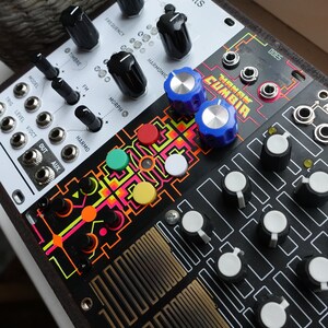 以下が含まれることがあります： カラフルなノブとボタンが付いた白黒のシンセサイザー。シンセサイザーは「TANGLED CUMBIA」というテキストのグラフィックデザインになっています。