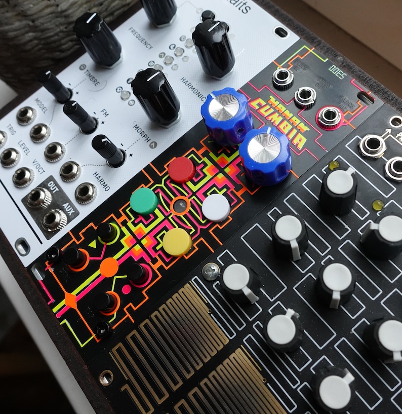 以下が含まれることがあります： カラフルなノブとボタンが付いた白黒のシンセサイザー。シンセサイザーは「TANGLED CUMBIA」というテキストのグラフィックデザインになっています。