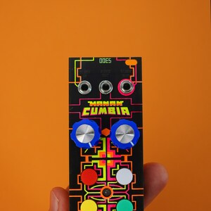以下が含まれることがあります： 「MANAN CUMBIA」と「ODES」の文字が印刷された黒と蛍光緑の電子音楽モジュール。モジュールには青いノブが2つ、赤いボタンが3つ、白いボタンが3つある。
