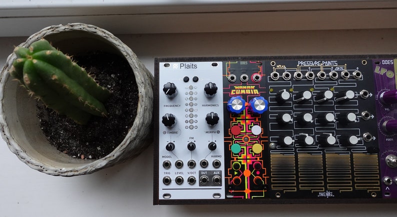 以下が含まれることがあります： 前面に「Plaits」と印刷された白黒のシンセサイザーモジュール。モジュールは、前面に「CUMBIA」と印刷された黒いシンセサイザーモジュールの隣にあります。モジュールは、赤、黄、緑、青のアクセントが付いたカラフルなグラフィックデザインです。モジュールは白い表面にあります。