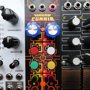 以下が含まれることがあります： 黒と白のシンセサイザーモジュールで、"HANAN CUMBIA"というテキストとカラフルなグラフィックデザインが施されています。モジュールにはノブ、ボタン、ジャックがあります。右上の隅には"ODES"というテキストが表示されています。