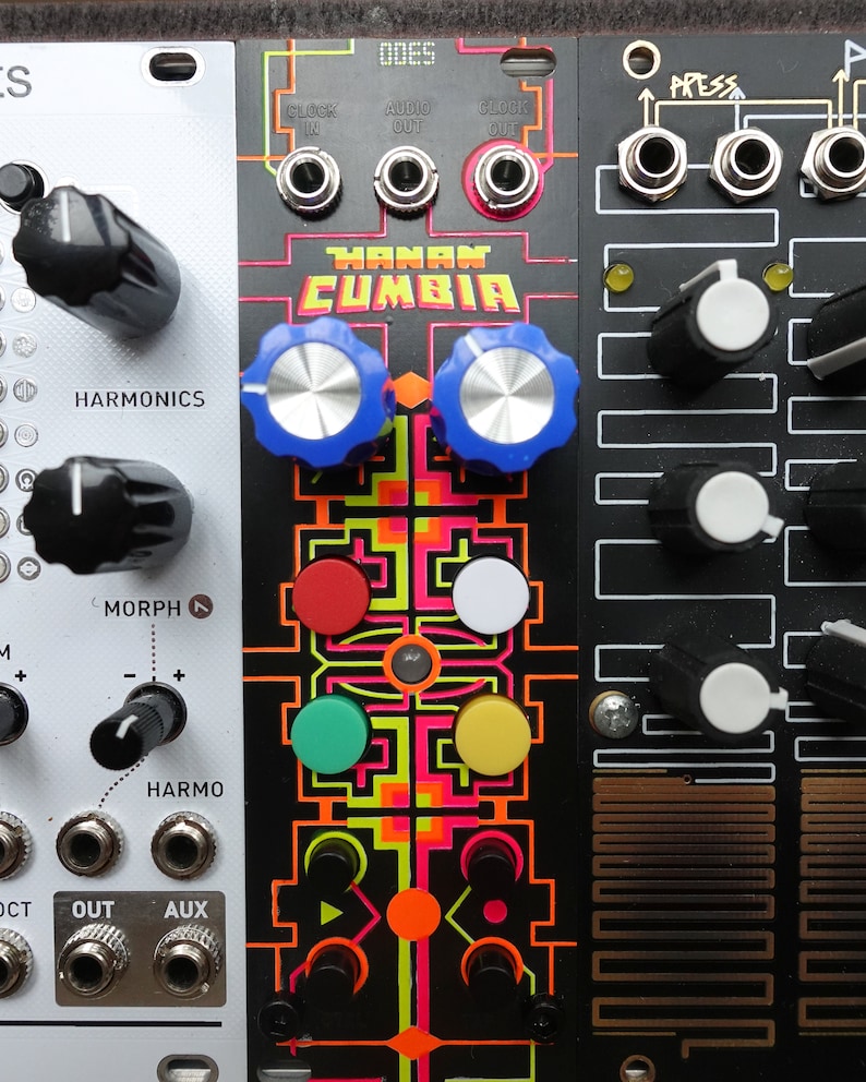 以下が含まれることがあります： 黒と白のシンセサイザーモジュールで、"HANAN CUMBIA"というテキストとカラフルなグラフィックデザインが施されています。モジュールにはノブ、ボタン、ジャックがあります。右上の隅には"ODES"というテキストが表示されています。