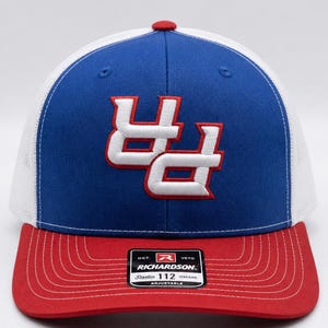 May include: A blue and white trucker hat with a red brim and a white embroidered logo. The hat features a blue front panel with a white embroidered logo and a white mesh back. The brim is red, and the hat is adjustable.