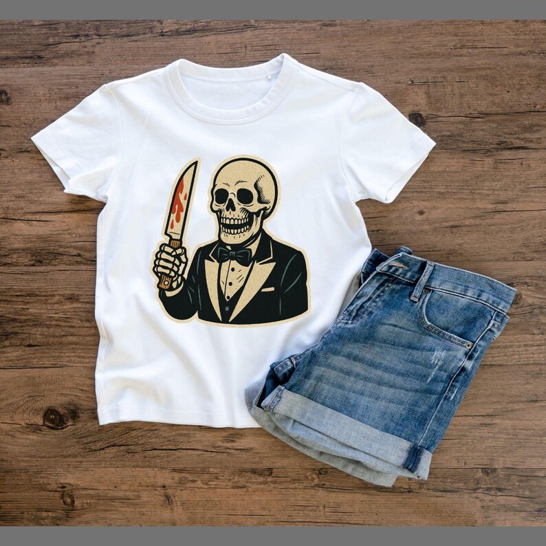 May include: White t-shirt featuring a cartoon skeleton in a tuxedo holding a bloody knife. The skeleton has a bow tie and is wearing a black tuxedo jacket. Blue denim shorts are next to the shirt on a wooden surface.