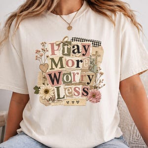 May include: Cream-colored t-shirt with the words "Pray More Worry Less" in a collage design. The design includes floral elements, a cross, and a heart. The shirt is a relaxed fit.