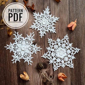 Può includere: Tre fiocchi di neve all'uncinetto bianchi su uno sfondo di legno marrone. Il testo "PATTERN PDF" è in un cerchio bianco nella parte superiore dell'immagine.