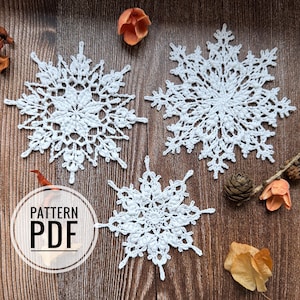 Op de afbeelding: Drie witte gehaakte sneeuwvlokken op een bruin houten oppervlak. De sneeuwvlokken zijn van verschillende grootte en hebben ingewikkelde patronen. De tekst "PATTERN PDF" is in wit op een zwarte cirkel geschreven.