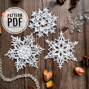 Może przedstawiać: Trzy białe, szydełkowe płatki śniegu na drewnianym tle. Płatki śniegu są delikatne i misterne, z różnymi wzorami. Tekst "PATTERN PDF" jest widoczny w lewym górnym rogu obrazu.