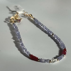 May include: A delicate gold bracelet with a mix of light blue, red, and clear gemstones. The bracelet has a lobster clasp closure.