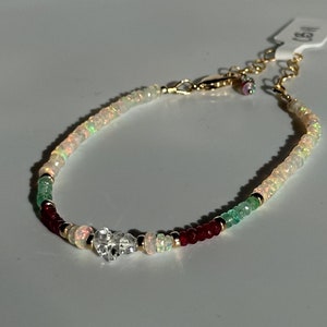 May include: Delicate bracelet featuring a mix of opal, emerald, and ruby beads, accented with gold-colored metal. A clear, faceted charm adds a touch of sparkle. The bracelet is adjustable and has a tag attached.