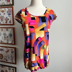 May include: A brightly coloured patterned top in shades of orange, pink, yellow, purple, and black. The short-sleeved shirt has a scoop neck and a ruffled hem. The abstract design is displayed on a mannequin.