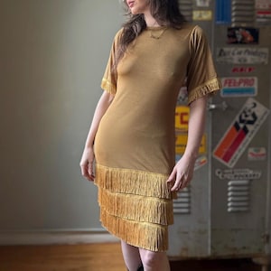 May include: A gold-colored dress with short sleeves and fringe detailing. The dress has a knee-length hemline and is paired with black cowboy boots. The dress has fringe on the sleeves and the hemline.