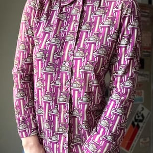 Puede incluir: Camisa abotonada de manga larga con un estampado repetido de coches antiguos en tonos morado, beige y blanco. La camisa presenta rayas verticales y un cuello clásico. Fabricada en tejido ligero.