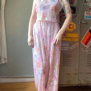 May include: A vintage two-piece pajama set in a light pink hue, adorned with a delicate floral pattern in shades of orange, blue, and pink. The top features a V-neckline with lace trim, and the pants have a drawstring waist. The set is made of a lightweight, flowing fabric.