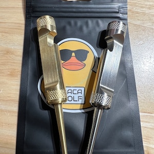 Puede incluir: Dos abridores de botellas con forma de tee de golf, uno dorado y otro plateado, con un logotipo de pato negro y amarillo en el embalaje. El logotipo dice "ACA GOLF".