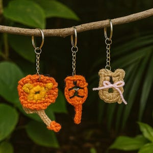 Puede incluir: Tres llaveros de ganchillo hechos a mano cuelgan de una rama. Los llaveros representan un león, un gato y un oso, cada uno con un diseño y una combinación de colores únicos. El león es naranja y amarillo, el gato es naranja y negro, y el oso es marrón con un lazo rosa.