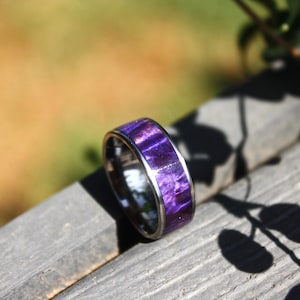 May include: A purple and silver ring with a wood inlay. The ring is on a wooden surface.
