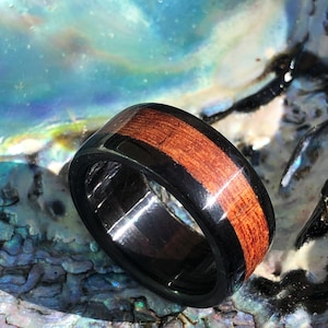 May include: A black and red wooden ring with a polished finish. The ring is made of two types of wood, one dark and one light. The ring is sitting on a blue and green iridescent surface.