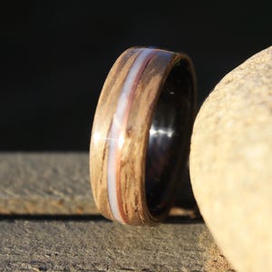 May include: A wooden ring with a white and copper inlay. The ring is set against a light brown background.