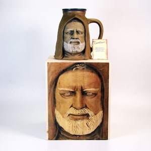 May include: A ceramic mug featuring a detailed portrait of a man with a beard and a hooded robe. The mug is displayed on a wooden box with a similar portrait of the man on the front. The box has a label that reads "Custom Creations".