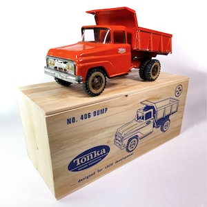 May include: A vintage red Tonka dump truck with a raised bed, sitting on a wooden box. The box features the text "NO. 406 DUMP" and a blue illustration of the truck. The truck has black tires and a silver front grill.
