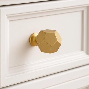 K&ouml;nnte beinhalten: Ein goldfarbener, geometrischer Schrankknauf mit facettiertem Design. Der Knauf ist an einer wei&szlig;en Schubladenfront montiert und zeigt ein modernes und dekoratives Hardware-Element. Die Form des Knaufs ist ein Dodekaeder.