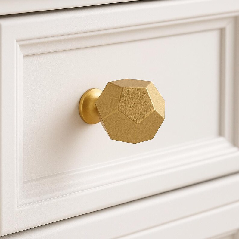 K&ouml;nnte beinhalten: Ein goldfarbener, geometrischer Schrankknauf mit facettiertem Design. Der Knauf ist an einer wei&szlig;en Schubladenfront montiert und zeigt ein modernes und dekoratives Hardware-Element. Die Form des Knaufs ist ein Dodekaeder.