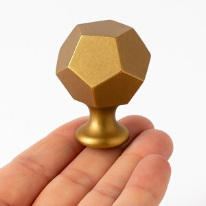 K&ouml;nnte beinhalten: Ein goldfarbener Schrankknauf mit einem geometrischen, vielseitigen Design. Der Knauf hat einen abgerundeten Sockel und eine zw&ouml;lffl&auml;chige Oberseite. Die Hardware wird in einer Hand vor wei&szlig;em Hintergrund gehalten.