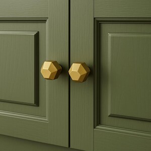 K&ouml;nnte beinhalten: Nahaufnahme von zwei goldenen geometrischen Schrankkn&ouml;pfen an einer gr&uuml;nen Schrankt&uuml;r. Die Kn&ouml;pfe haben ein facettiertes Design und ein metallisches Finish. Die Schrankt&uuml;r hat ein eingelassenes Paneeldesign.