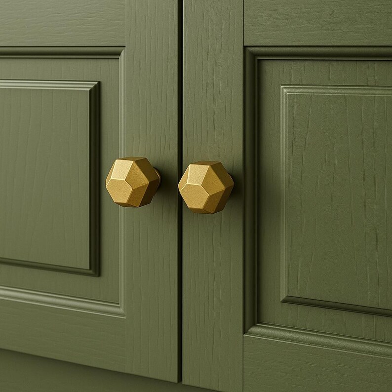 K&ouml;nnte beinhalten: Nahaufnahme von zwei goldenen geometrischen Schrankkn&ouml;pfen an einer gr&uuml;nen Schrankt&uuml;r. Die Kn&ouml;pfe haben ein facettiertes Design und ein metallisches Finish. Die Schrankt&uuml;r hat ein eingelassenes Paneeldesign.
