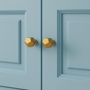 K&ouml;nnte beinhalten: Nahaufnahme von zwei goldenen Schrankkn&ouml;pfen mit geometrischem Design. Die Kn&ouml;pfe sind an einer hellblauen Schrankt&uuml;r befestigt. Die Kn&ouml;pfe haben eine matte Oberfl&auml;che und ein modernes Aussehen.