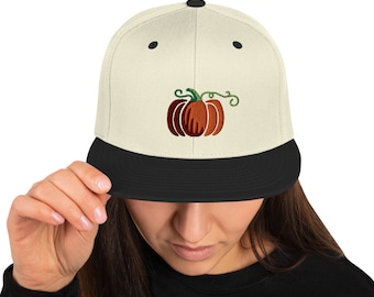 May include: A beige and black baseball cap with an embroidered pumpkin design. The pumpkin is orange with a green stem and leaves.