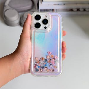 May include: Clear phone case with an iridescent, shimmering background. The case has a clear border and contains small, floating heart-shaped and character charms. The phone case is designed to fit a smartphone.