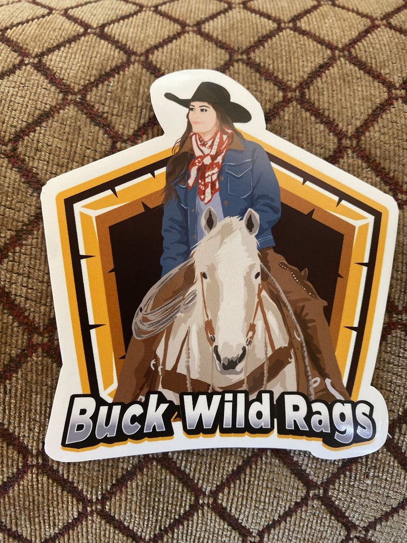 May include: A sticker featuring a cowgirl on a white horse, wearing a black hat and denim jacket. The sticker has a brown and orange border with the text "Buck Wild Rags" in a bold, stylized font.