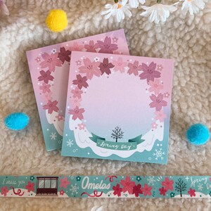 May include: Two pink and white floral patterned memo pads with the text "Spring Day" on the bottom of the larger pad. The pads are on a white fluffy surface.