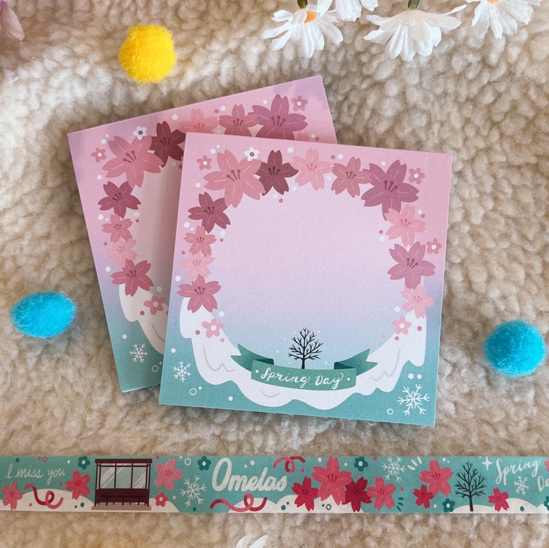 May include: Two pink and white floral patterned memo pads with the text "Spring Day" on the bottom of the larger pad. The pads are on a white fluffy surface.