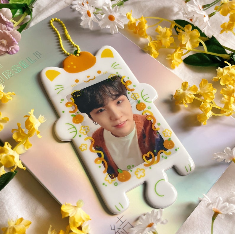May include: A white cat-shaped photo holder with a yellow strap and orange accents. The holder frames a photo of a person. The holder is decorated with orange fruit and green leaves. Yellow flowers and daisies surround the holder.