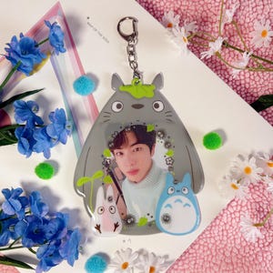 Puede incluir: Un llavero Totoro gris con un inserto de foto transparente que presenta a un hombre con cabello oscuro. El inserto de foto está rodeado de hojas verdes y pequeños personajes Totoro negros y blancos. El llavero tiene una cadena plateada y una hoja verde en la parte superior de la cabeza de Totoro.