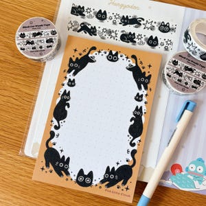 May include: A rectangular notepad with a tan border and a white dotted center featuring black cat illustrations. Two rolls of black cat washi tape, a sheet of washi tape with cat patterns, and a light blue pen are arranged around the notepad.