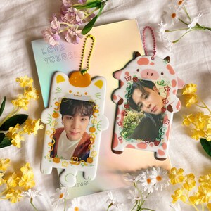 May include: Two decorative photo holders, one cat-shaped and one cow-shaped, each holding a photograph of a person. The cat holder is white with orange accents, and the cow holder is pink and white. Both have keychains. The background includes flowers and a book with the text "LOVE YOURSELF".