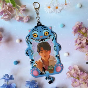 Op de afbeelding: Een blauwe tijgervormige sleutelhanger met een foto van een persoon erin. De sleutelhanger heeft een zilveren sluiting en is versierd met roze en zwarte accenten. De tijger heeft een lachend gezicht en is omringd door bloemen.