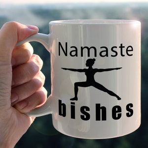 May include: White ceramic mug with the words "Namaste bishes" in black lettering. A black silhouette of a person in a yoga pose is centered on the mug. The mug is held by a hand, with a blurred background.