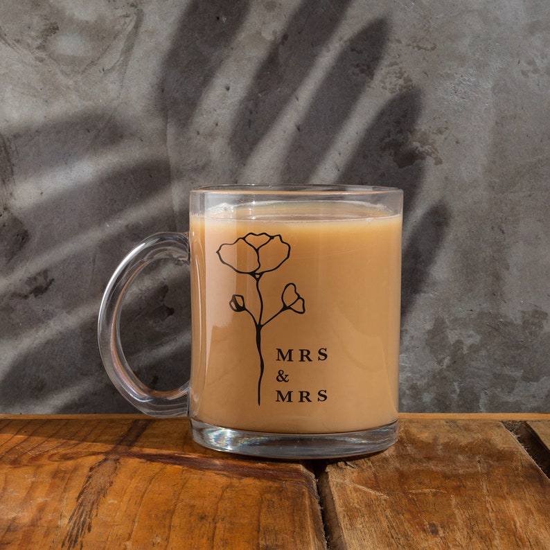 May include: A clear glass mug with a black floral design and the text "MRS & MRS" on the side. The mug is filled with a light brown beverage.