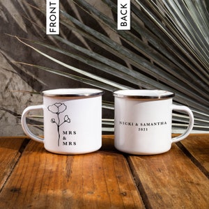 May include: Two white enamel mugs with black text and a simple floral design. One mug says "MRS & MRS" and the other says "NICKI & SAMANTHA 2021".