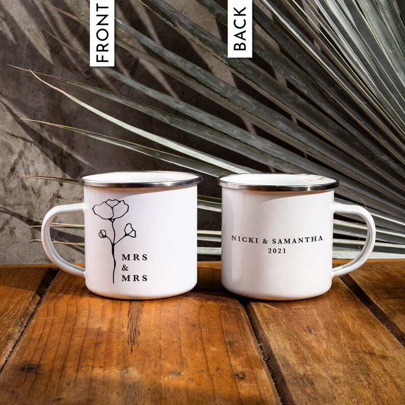 May include: Two white enamel mugs with black text and a simple floral design. One mug says "MRS & MRS" and the other says "NICKI & SAMANTHA 2021".