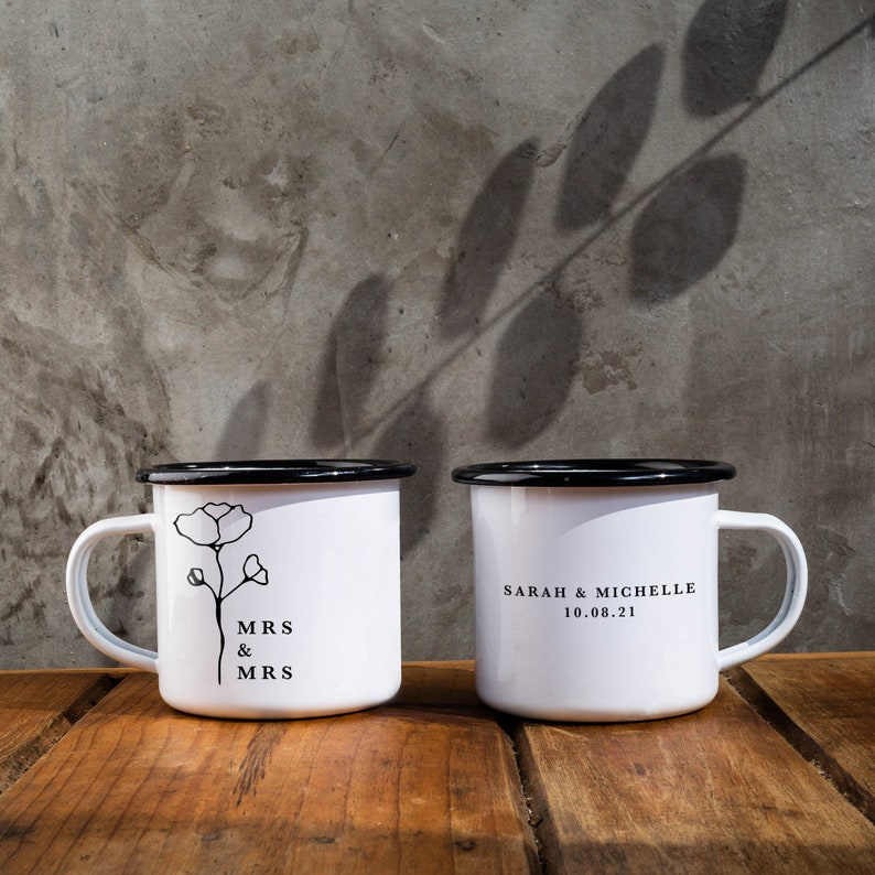 May include: Two white enamel mugs with black rims. One mug has a floral design and the text "MRS & MRS". The other mug has the text "SARAH & MICHELLE 10.08.21".
