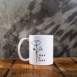 May include: White ceramic mug with a black outline of a flower and the text "MRS & MRS" printed on the front.
