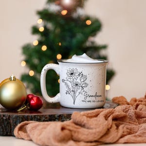 Może przedstawiać: Biały kubek ceramiczny z czarnym wzorem kwiatowym i tekstem "Grandma You Are So Loved". Kubek jest wypełniony białym napojem i stoi na drewnianej powierzchni z brązową tkaniną w tle.