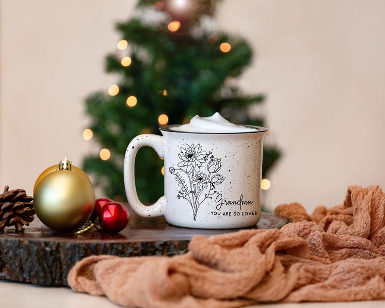 Może przedstawiać: Biały kubek ceramiczny z czarnym wzorem kwiatowym i tekstem "Grandma You Are So Loved". Kubek jest wypełniony białym napojem i stoi na drewnianej powierzchni z brązową tkaniną w tle.